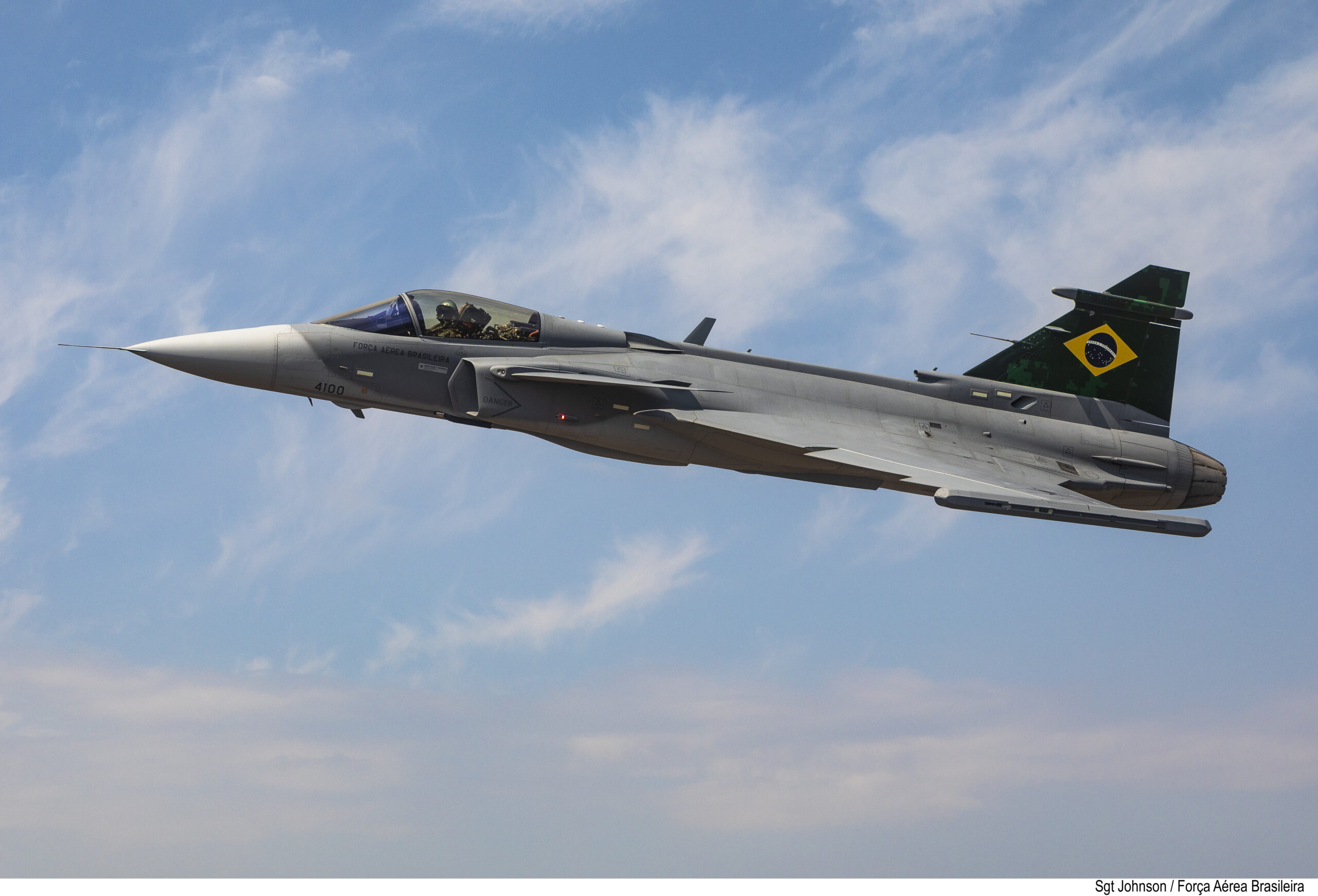 Avião Gripen em voô, com a frente para a esquerda da foto. É uma aeronave cinza, de bico afiado, com a bandeira do Brasil na cauda. Por trás, um ceu azul claro e poucas nuvens.